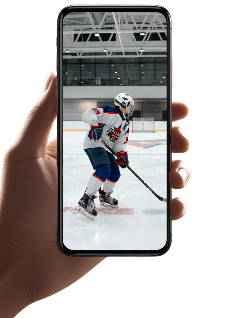 Person holding smartphone capturing a photo of a young hockey player on ice in an indoor rink.