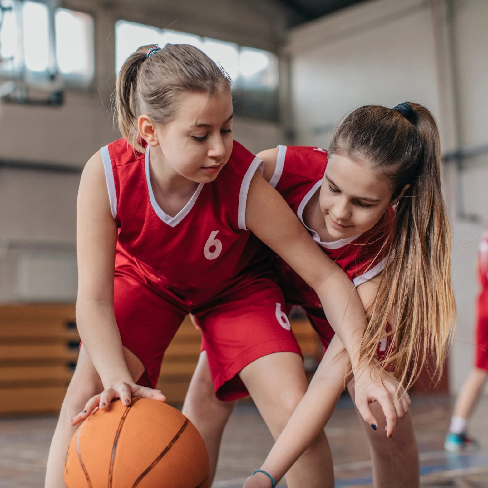 Two young girls in red basketball uniforms playing basketball in an indoor gym, vying for possession of the ball.