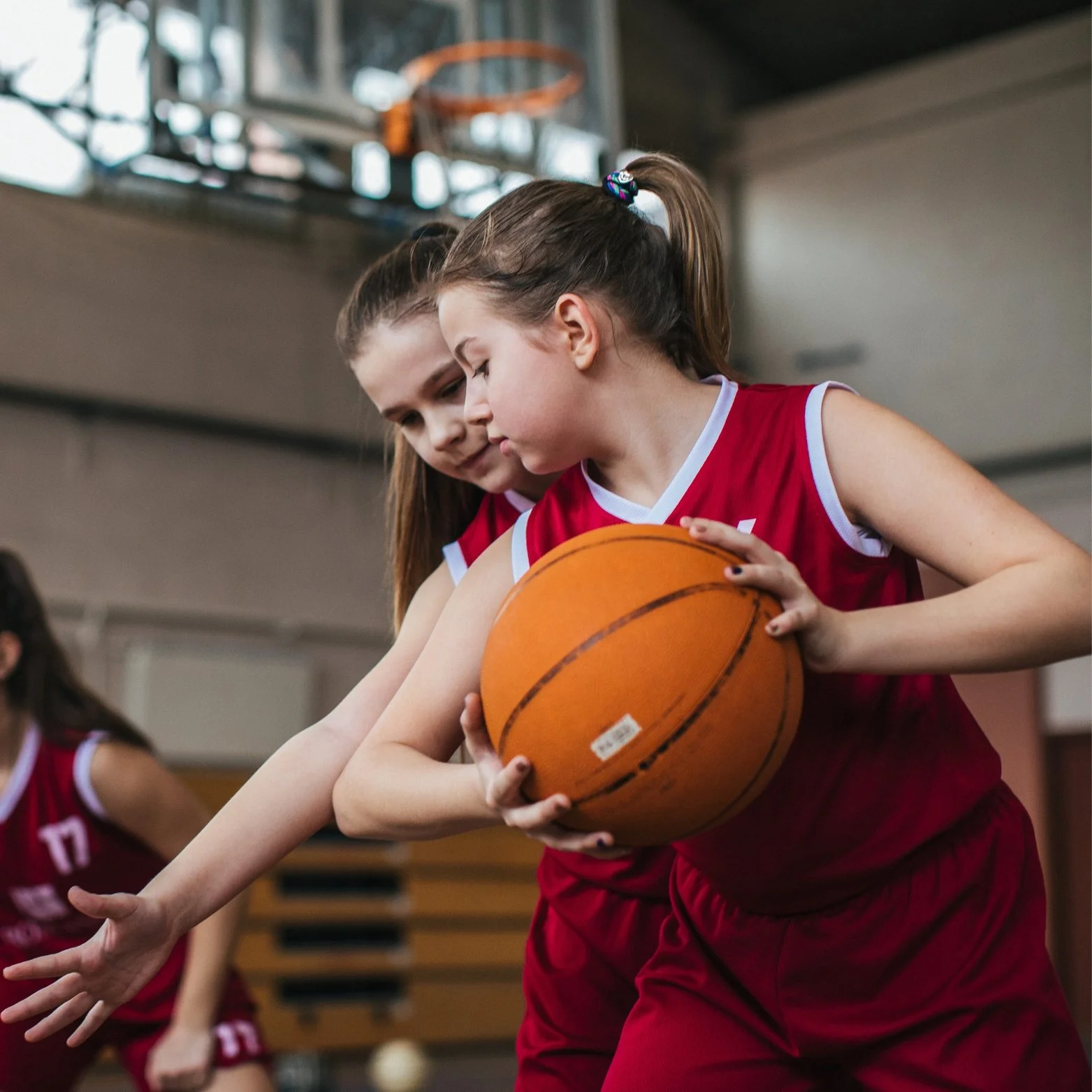 Two young girls in red basketball uniforms playing in an indoor gym, one holding a basketball.