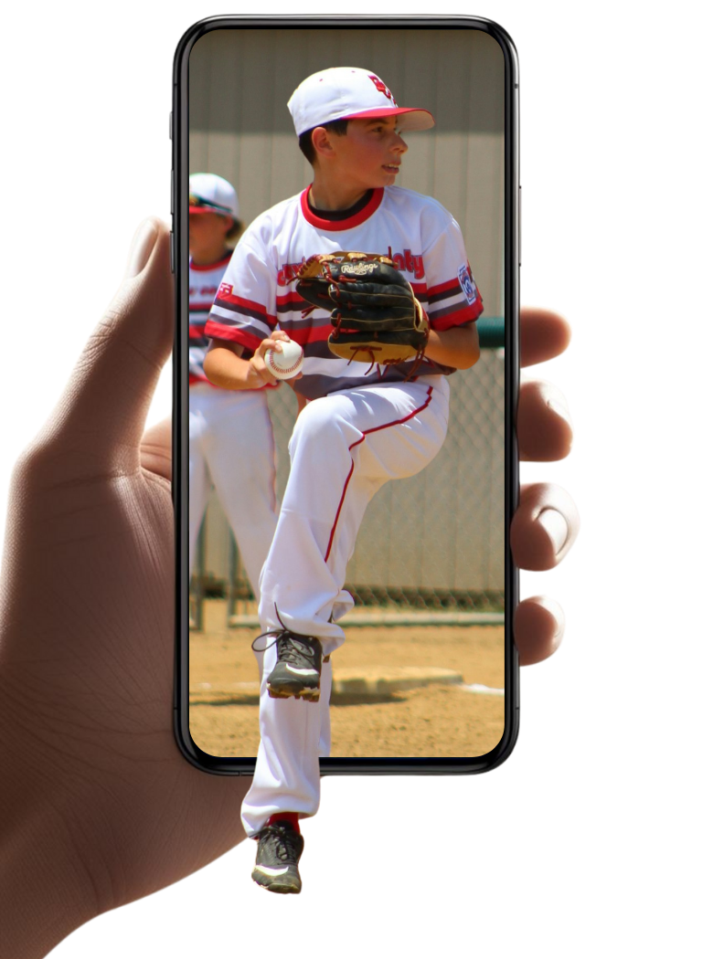 A person holding a smartphone with a photo of a young boy pitching a baseball during a game, wearing a white baseball uniform and cap.