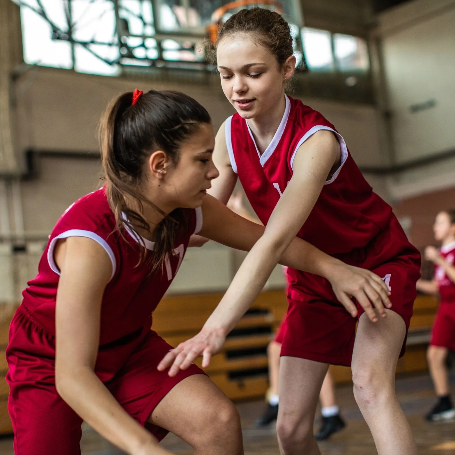Two young female basketball players in red uniforms play a game inside a gymnasium, with one girl attempting to guard the other.
