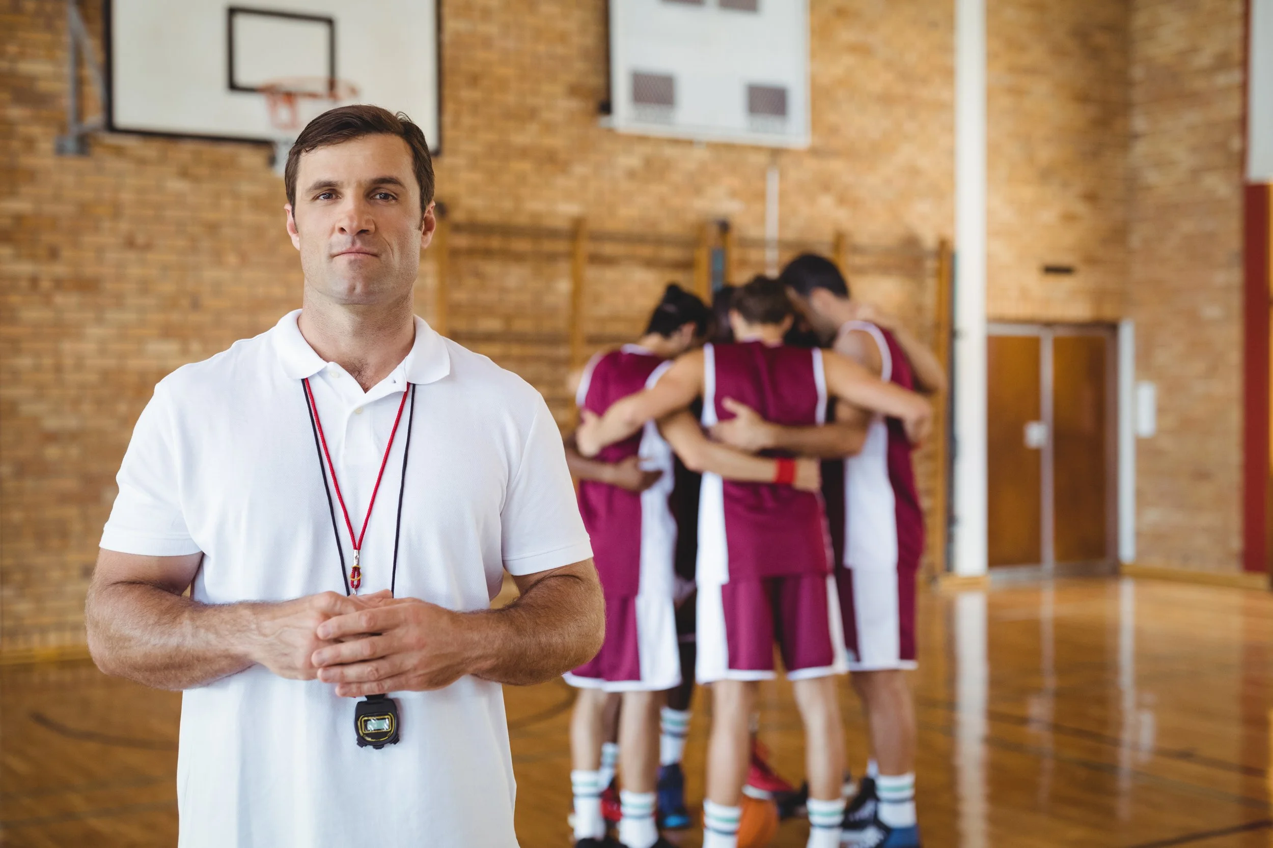 Basketball coach standing in front of a team of players huddled together in a gymnasium.