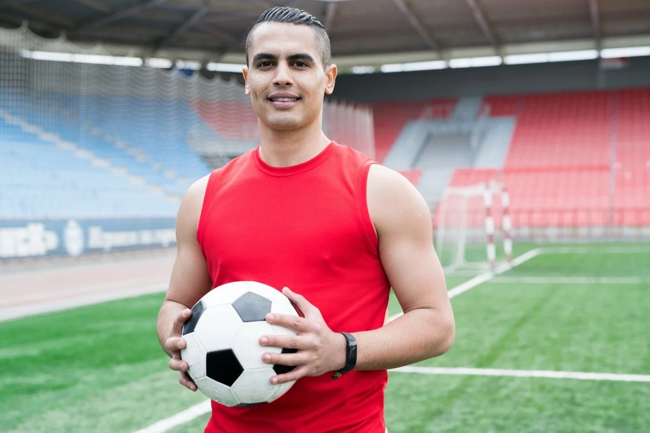 Young man in a red sleeveless shirt holding a soccer ball on a soccer field with empty stadium seating in the background.
