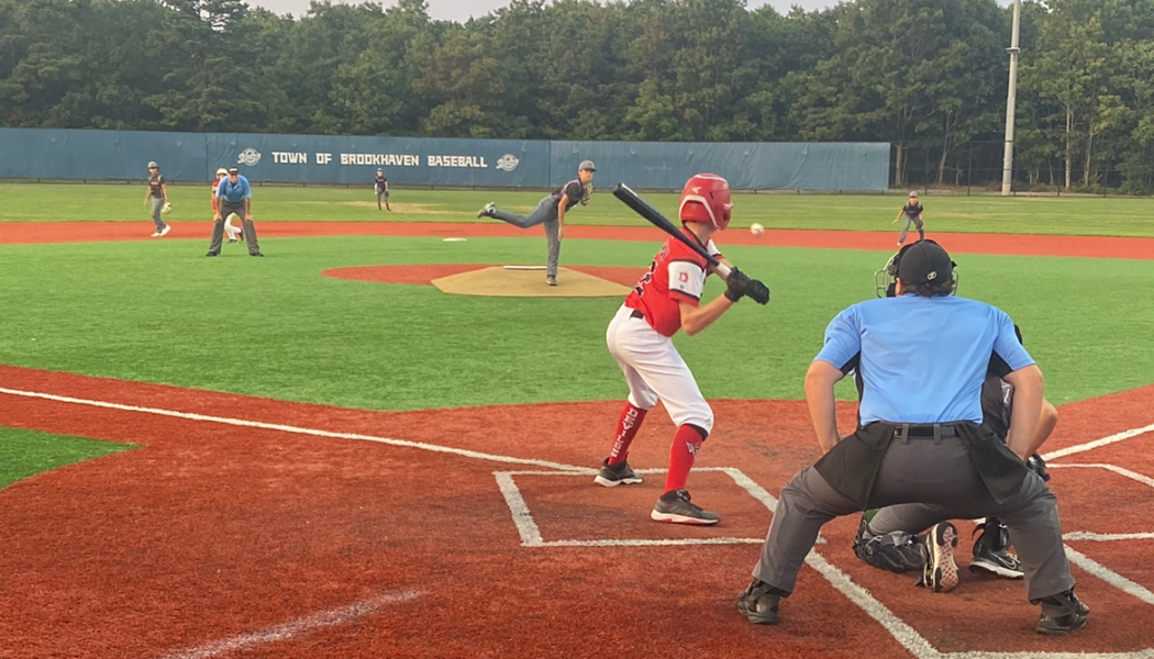 A baseball player in a purple and black uniform swings at a pitched ball during a game on the field, with an umpire and catcher observing.