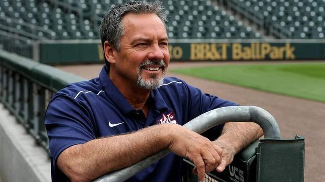 A middle-aged man with gray hair and a beard smiling at the camera, wearing a navy blue polo shirt with UCONN embroidered on it, outdoors with a blurred background.