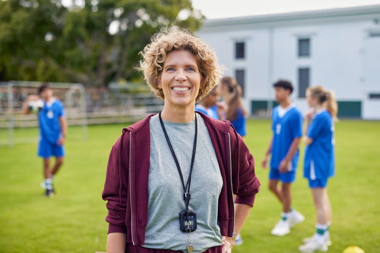 A smiling female coach with short curly blond hair standing on a soccer field in front of young players in blue uniforms, with a white building and trees in the background.