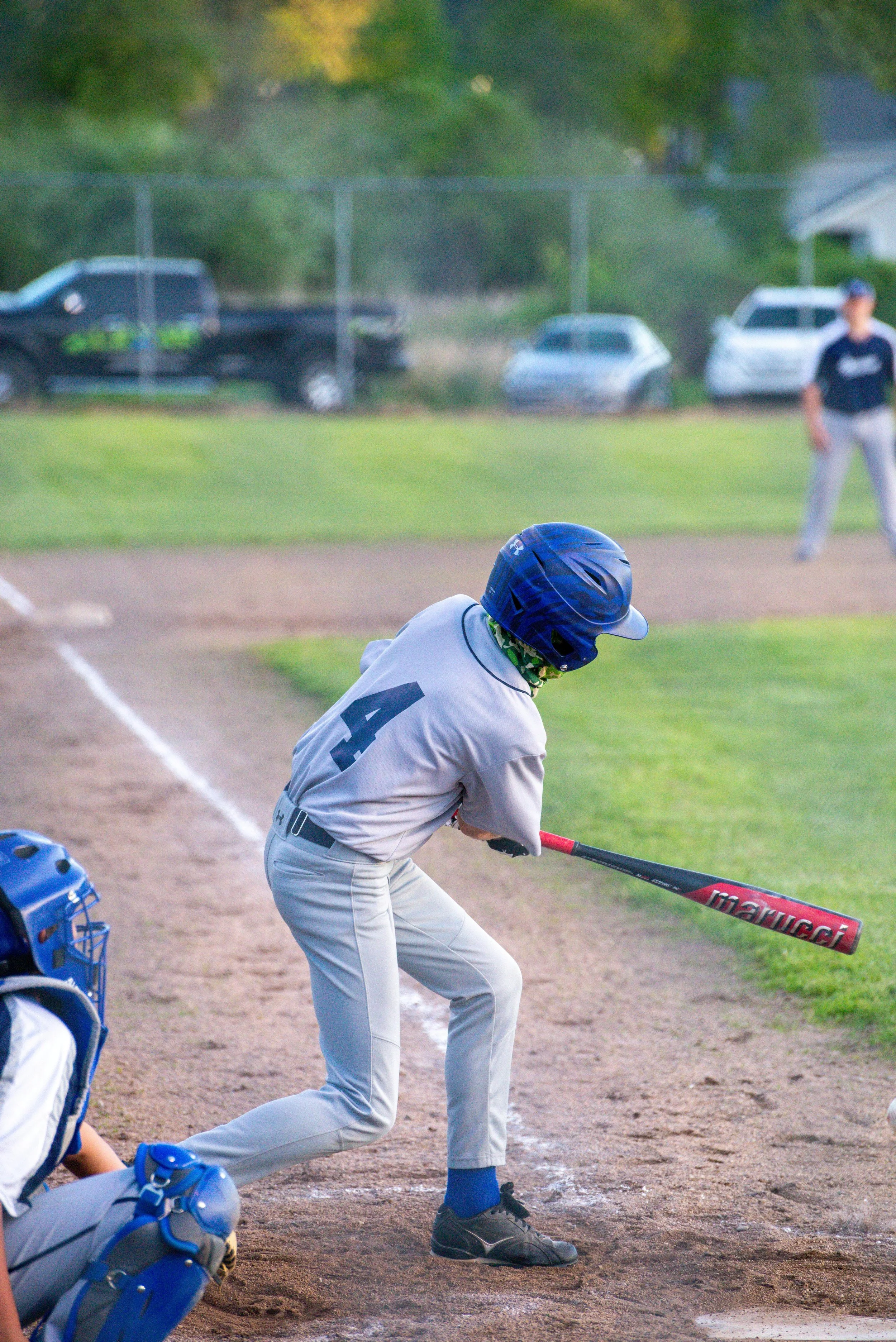 A young softball batter wearing a black helmet, orange jersey, and black pants preparing to hit a ball on a dirt field during a game, with two outfield players and green trees in the background.
