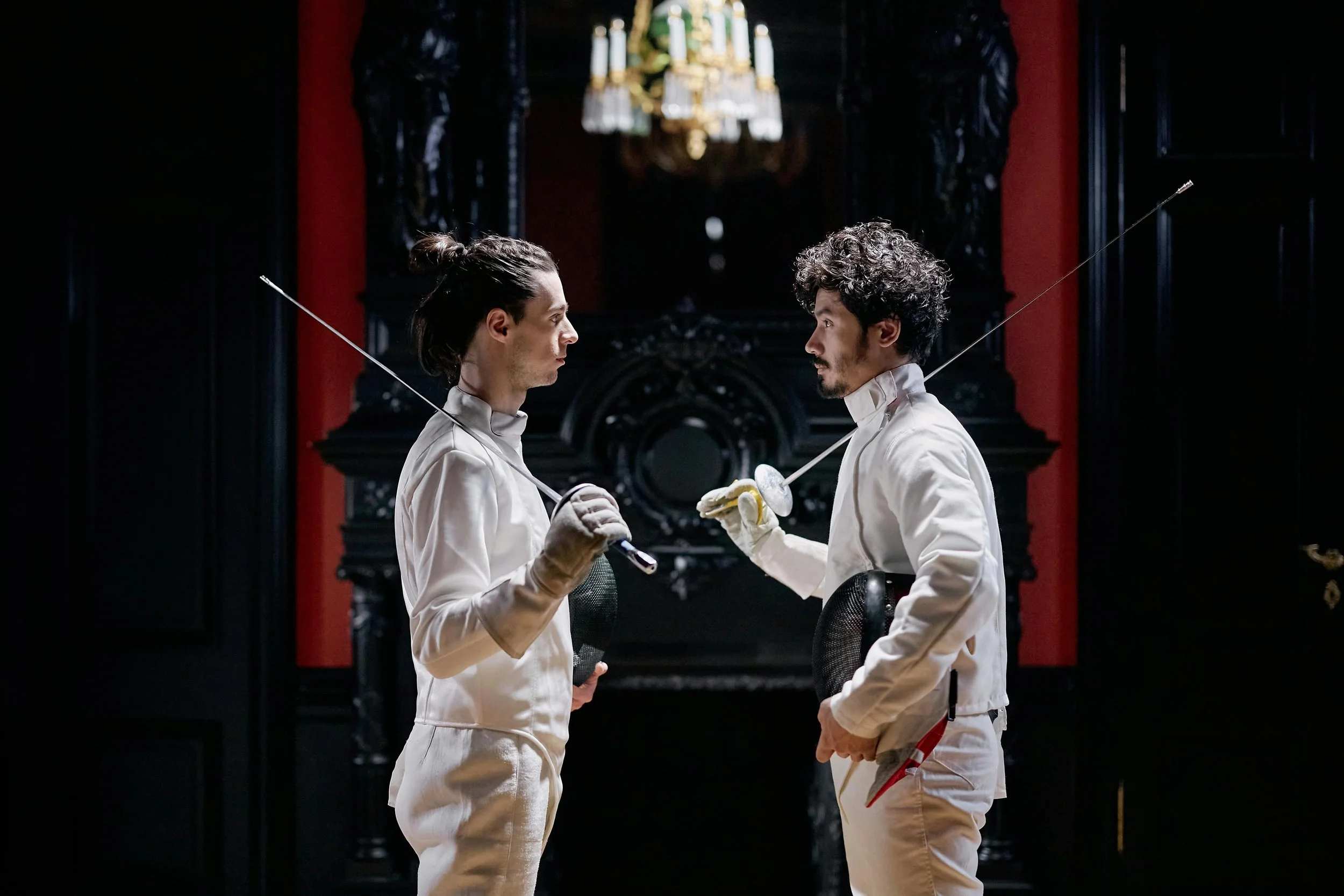 Two men dressed in white fencing uniforms face each other in a dimly lit room with black and red walls, holding fencing masks and swords, with an ornate chandelier hanging overhead.