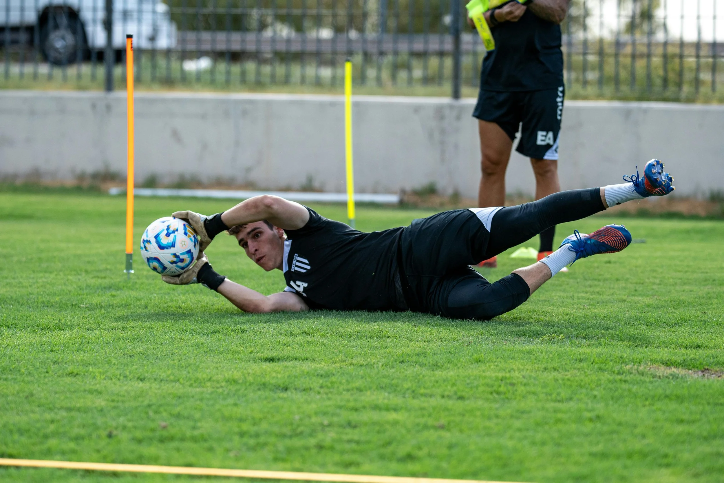 A soccer goalkeeper is diving on the grass to catch a football during training, with a coach standing nearby.