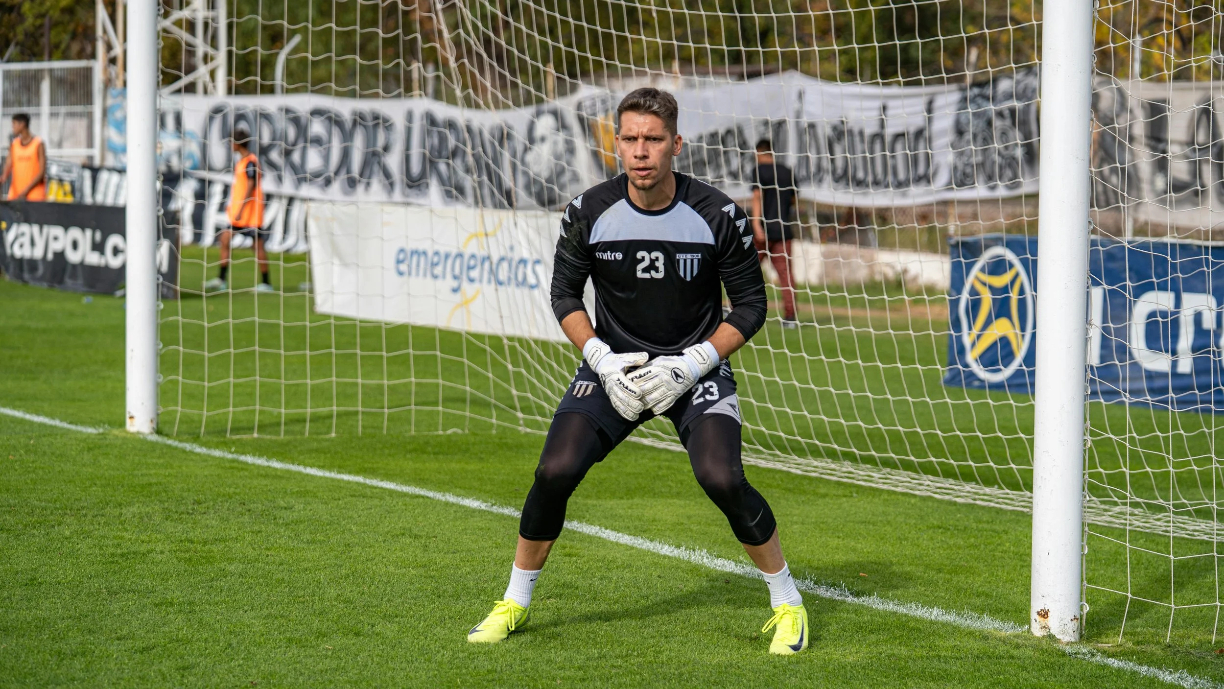 A male soccer goalkeeper in black uniform with the number 23 stands in front of the goal on a grass field, wearing white gloves and bright yellow cleats, with a focused expression.
