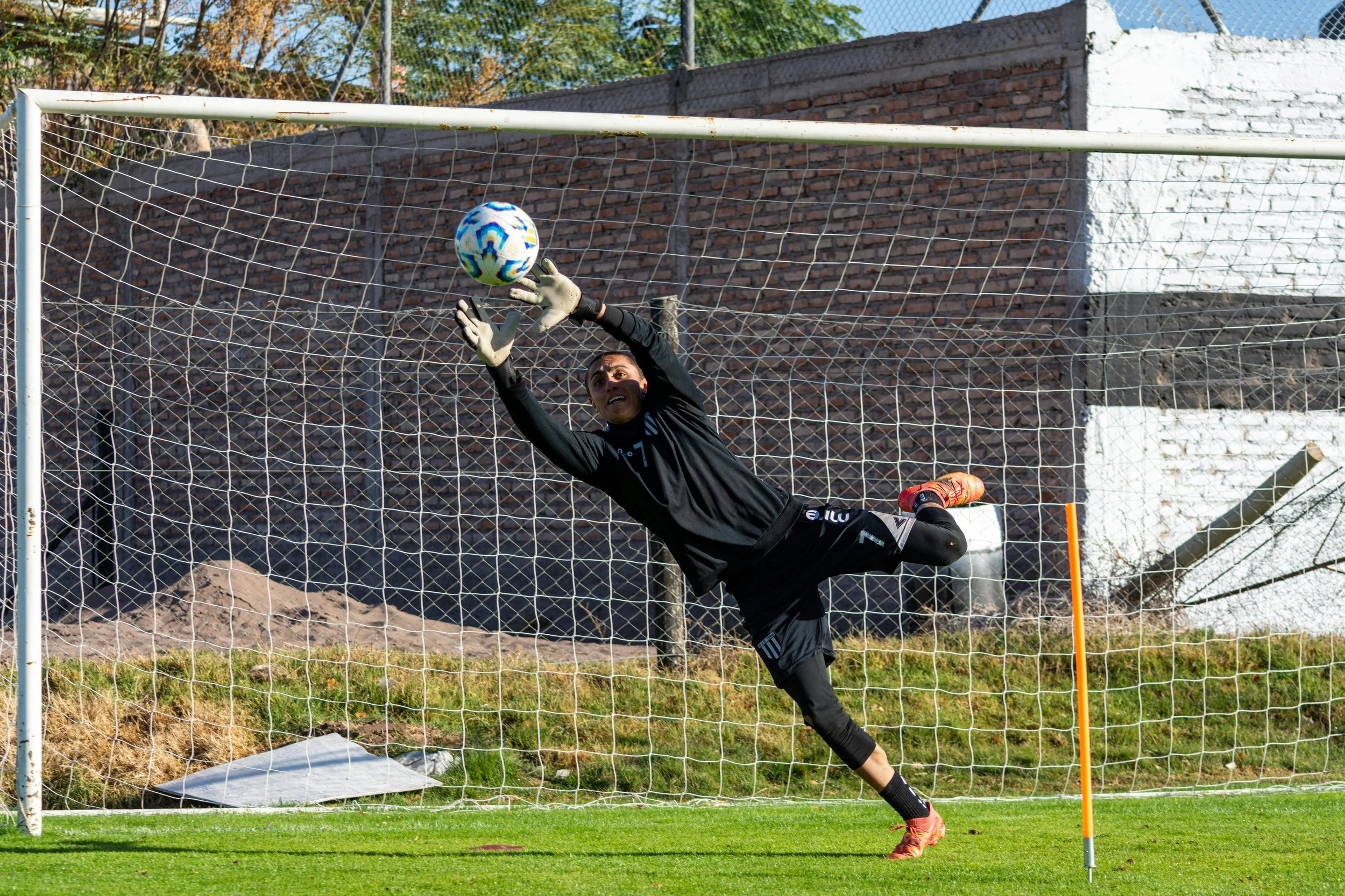A soccer goalkeeper in a black uniform is jumping to save a ball near the goalpost on a grassy field with a brick wall and a building in the background.
