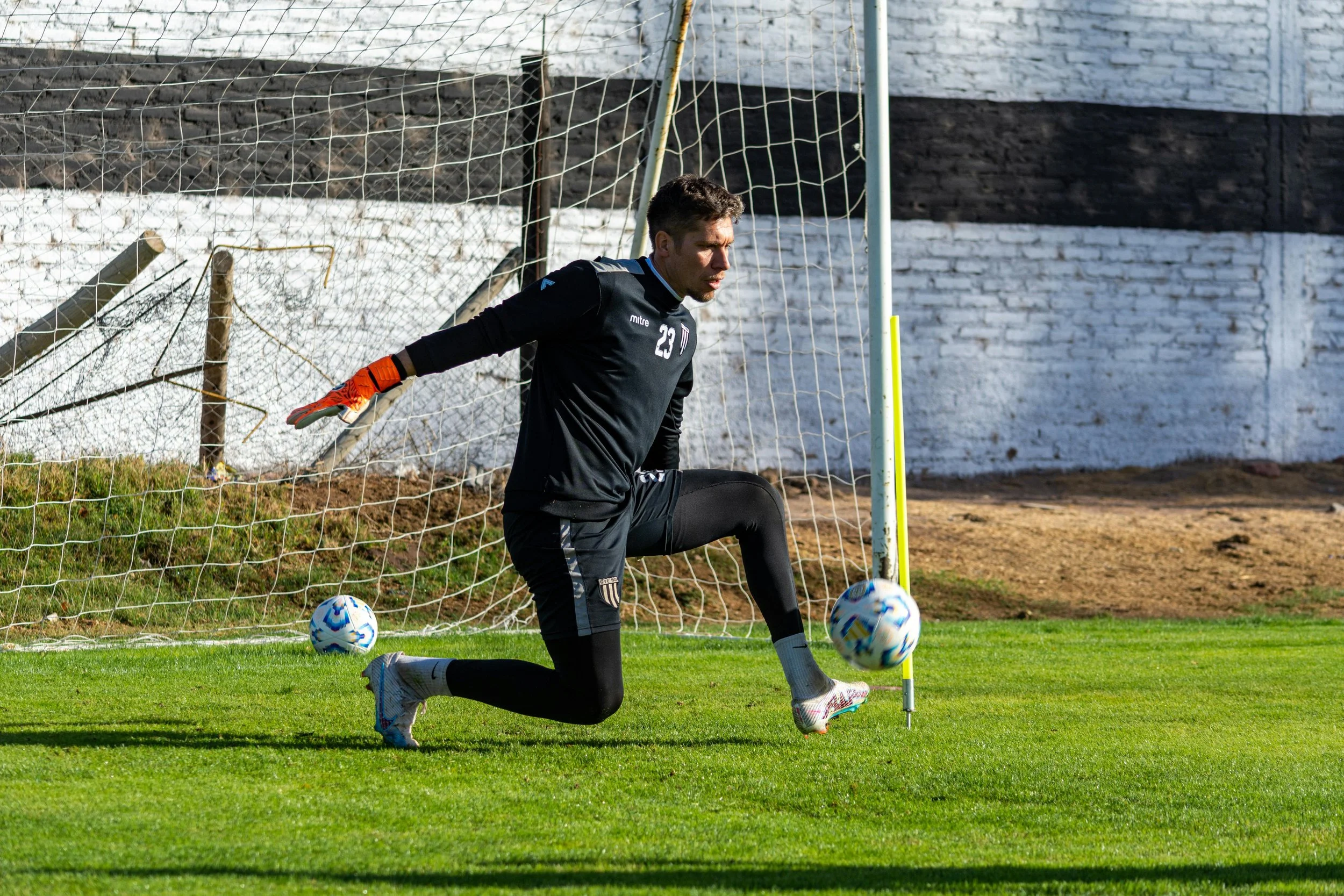 A soccer goalkeeper in black uniform and gloves practicing kicking a soccer ball outside near the goalpost on a grassy field.