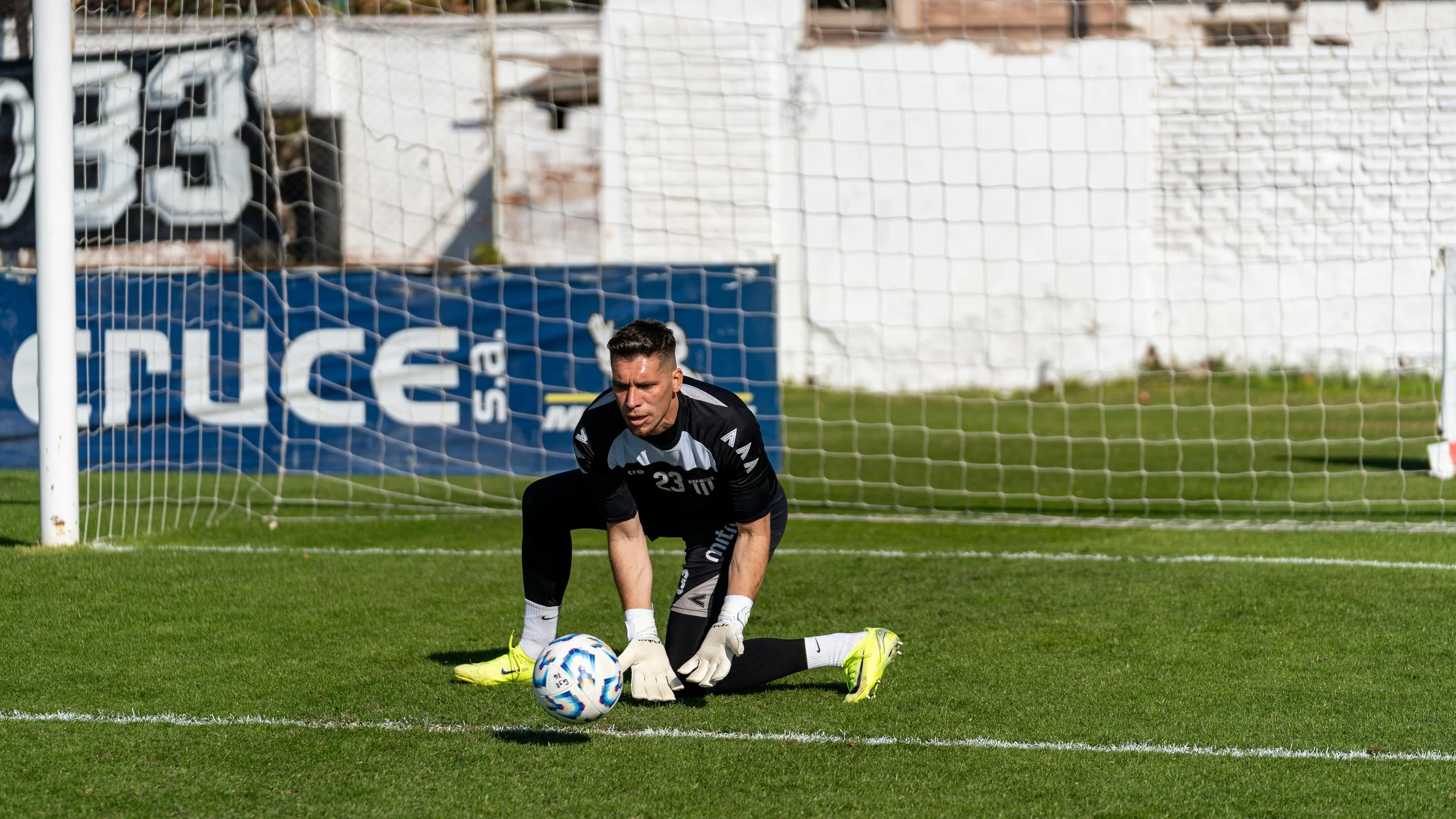 A soccer goalkeeper in a black uniform kneeling on the grass, preparing to kick a soccer ball in front of the goal.
