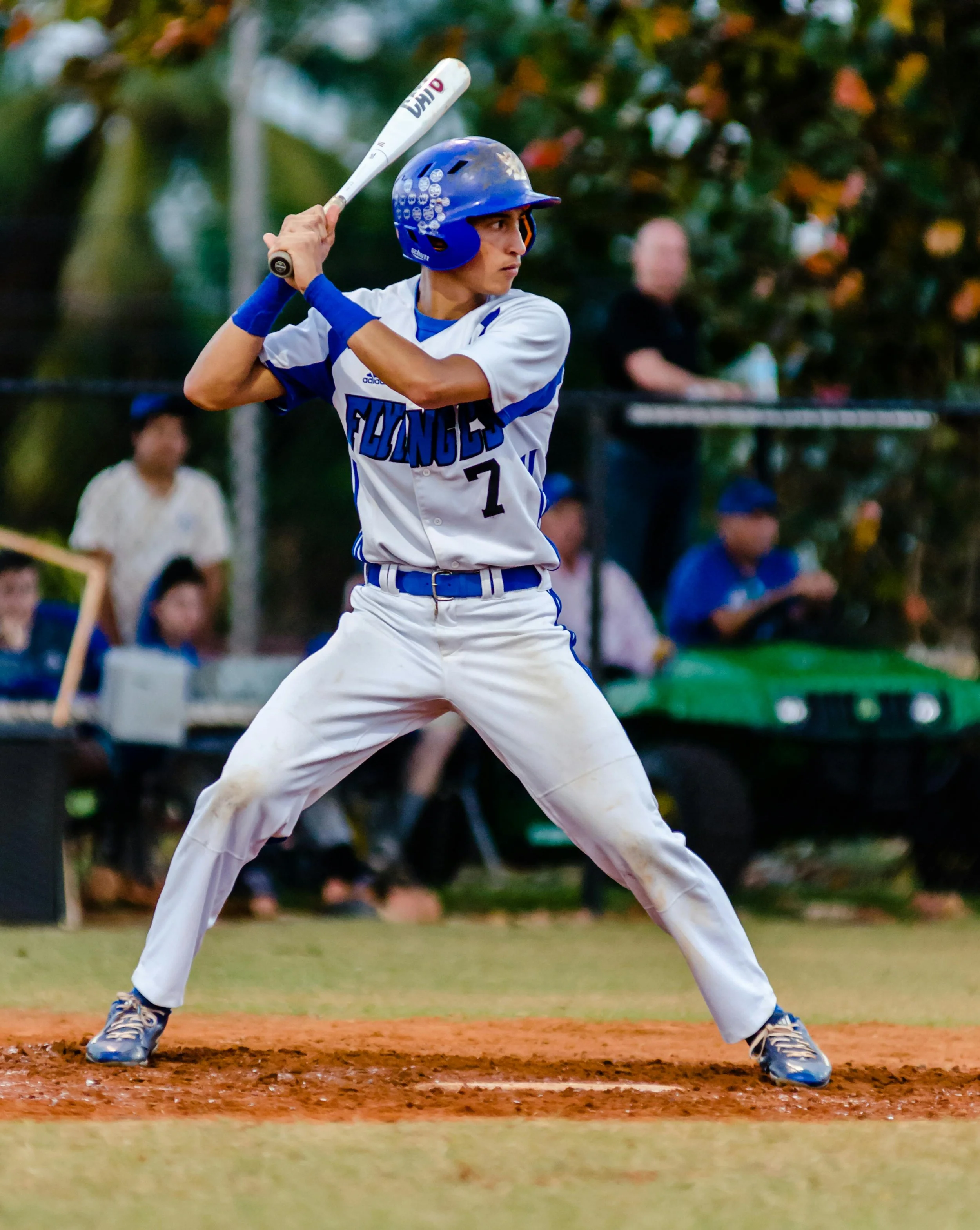 A young baseball player in a white and blue uniform, wearing a helmet, holds a bat in a batting stance on the field. Spectators sit in the background during a game at dusk.