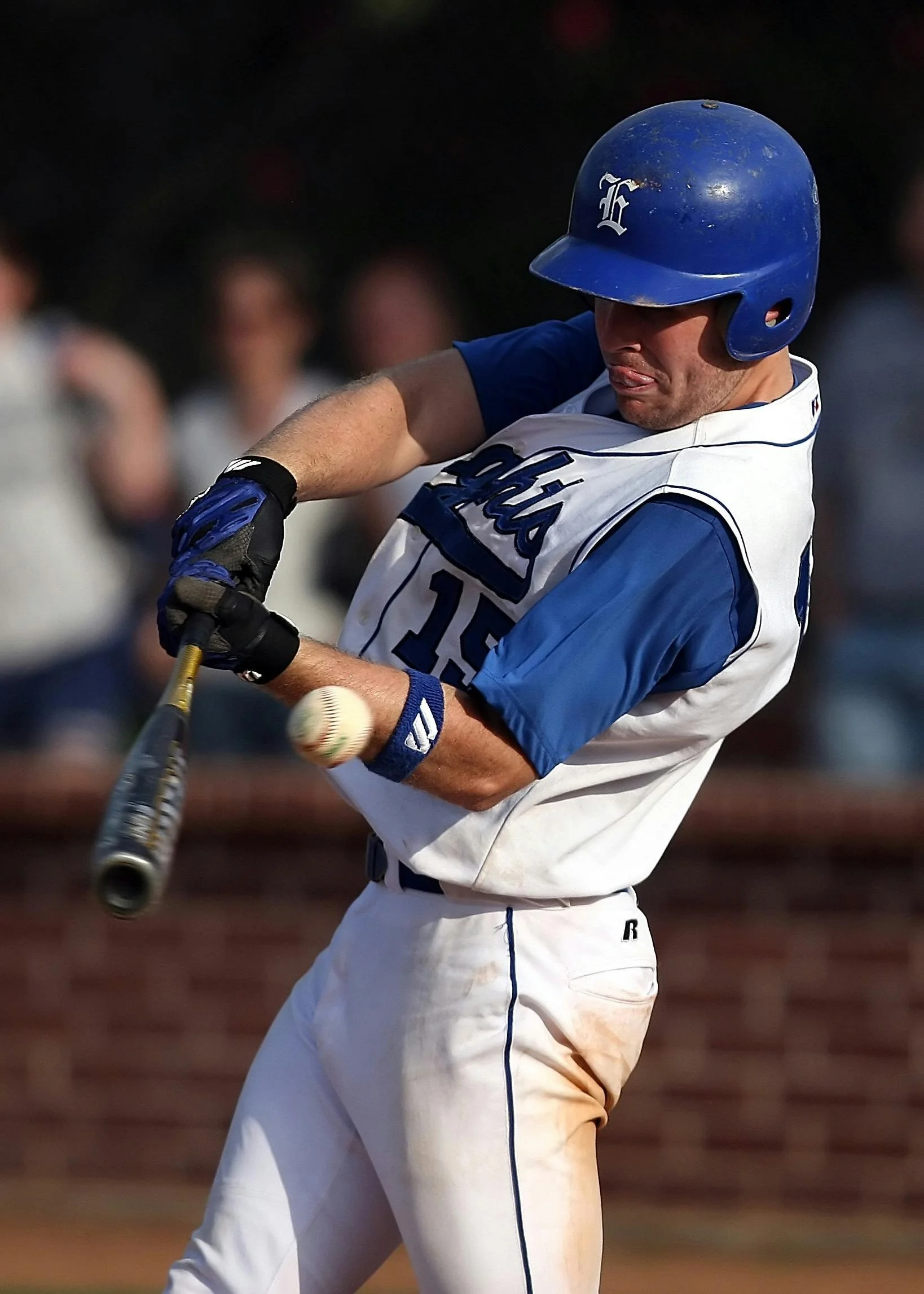 A baseball player wearing a blue helmet, white jersey with blue accents, and white pants is in the process of hitting a baseball with a bat during a game. The background shows spectators.