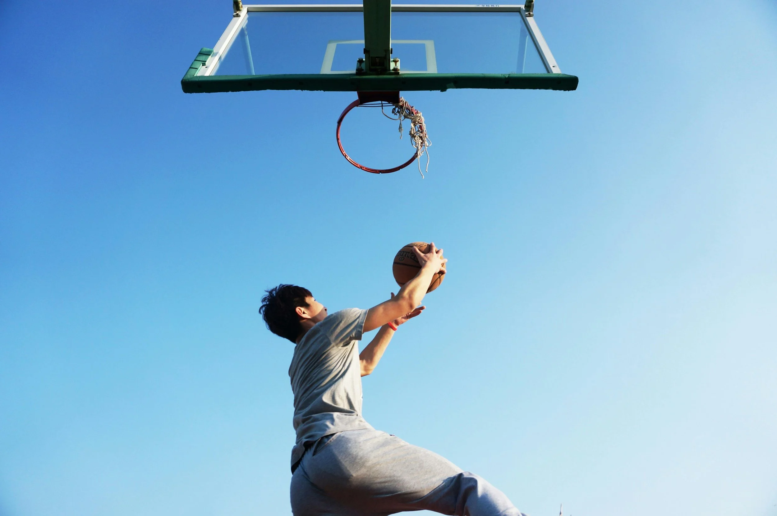 Person playing basketball outdoors, jumping to make a shot toward the hoop, against a clear blue sky.