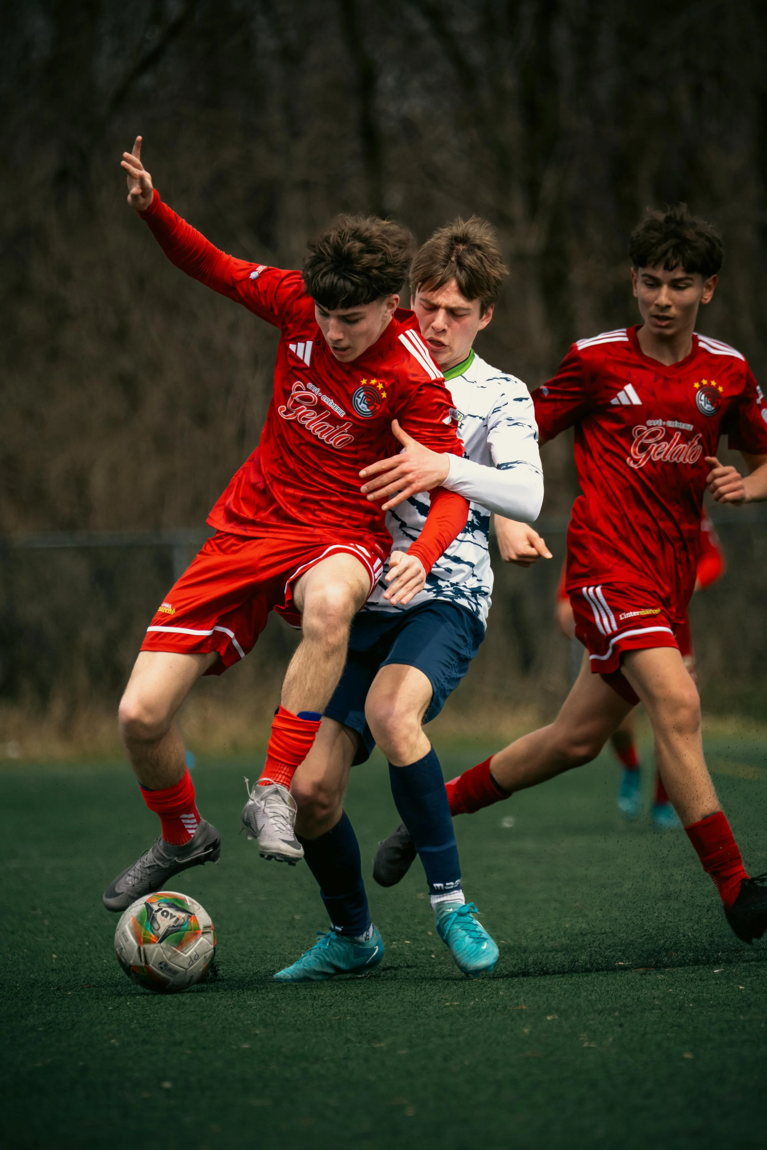 Three young male soccer players competing for possession of the ball during a match, with two players in red uniforms and one in a white and blue uniform, on a grassy field with trees in the background.