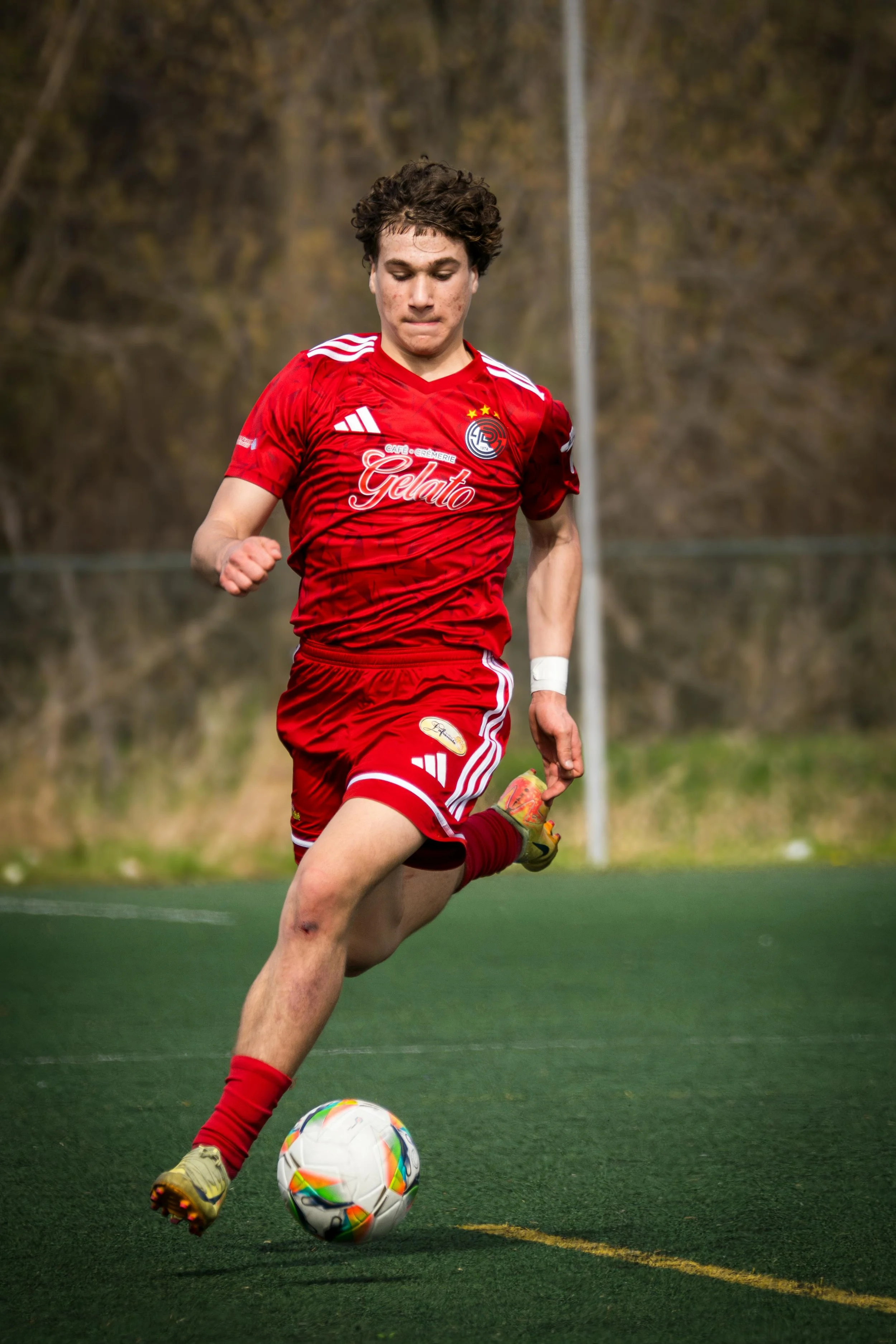 A young male soccer player in a red uniform kicking a soccer ball on an outdoor field.