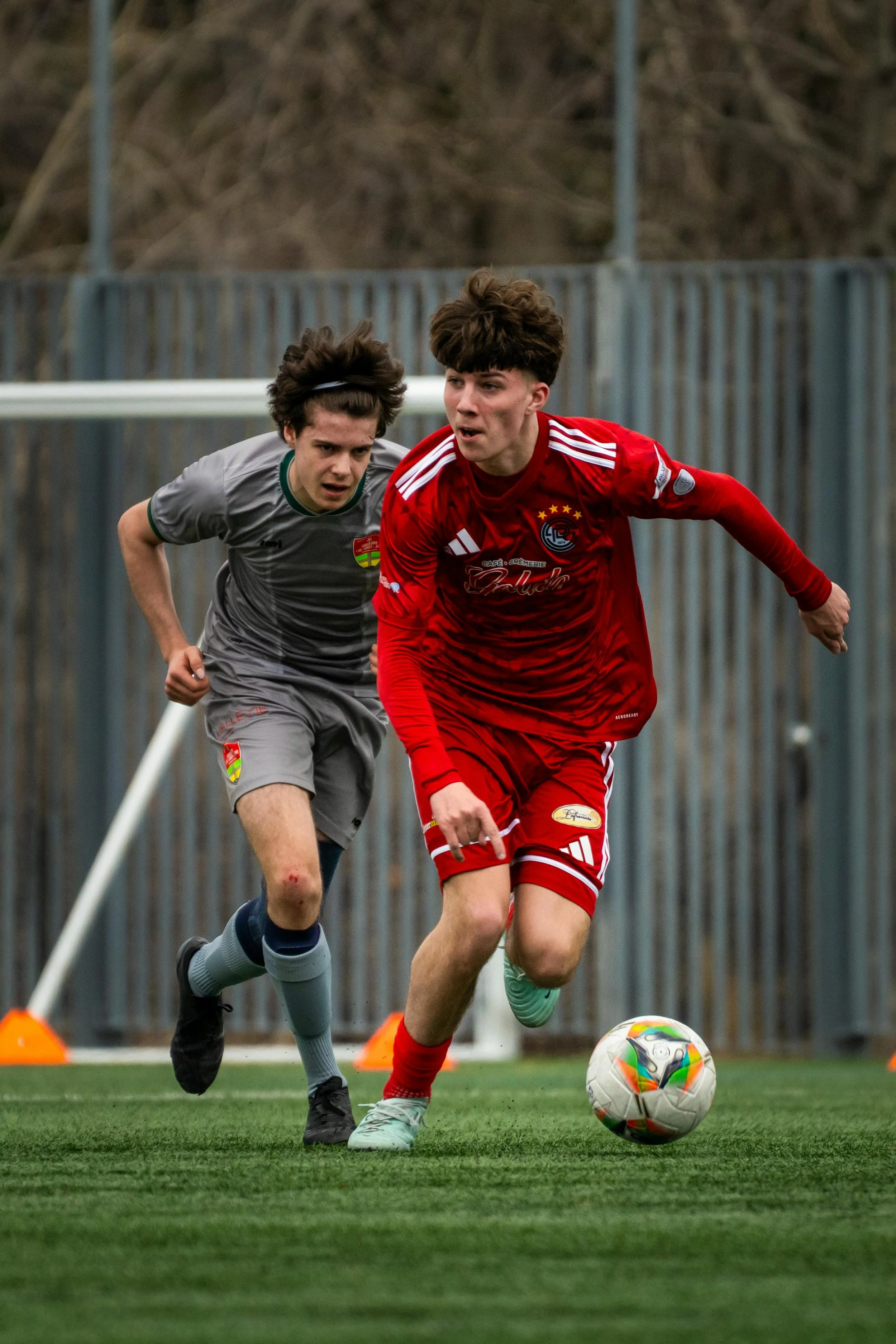 Two young soccer players competing for control of the ball on a field, with one wearing a red uniform and the other in gray.