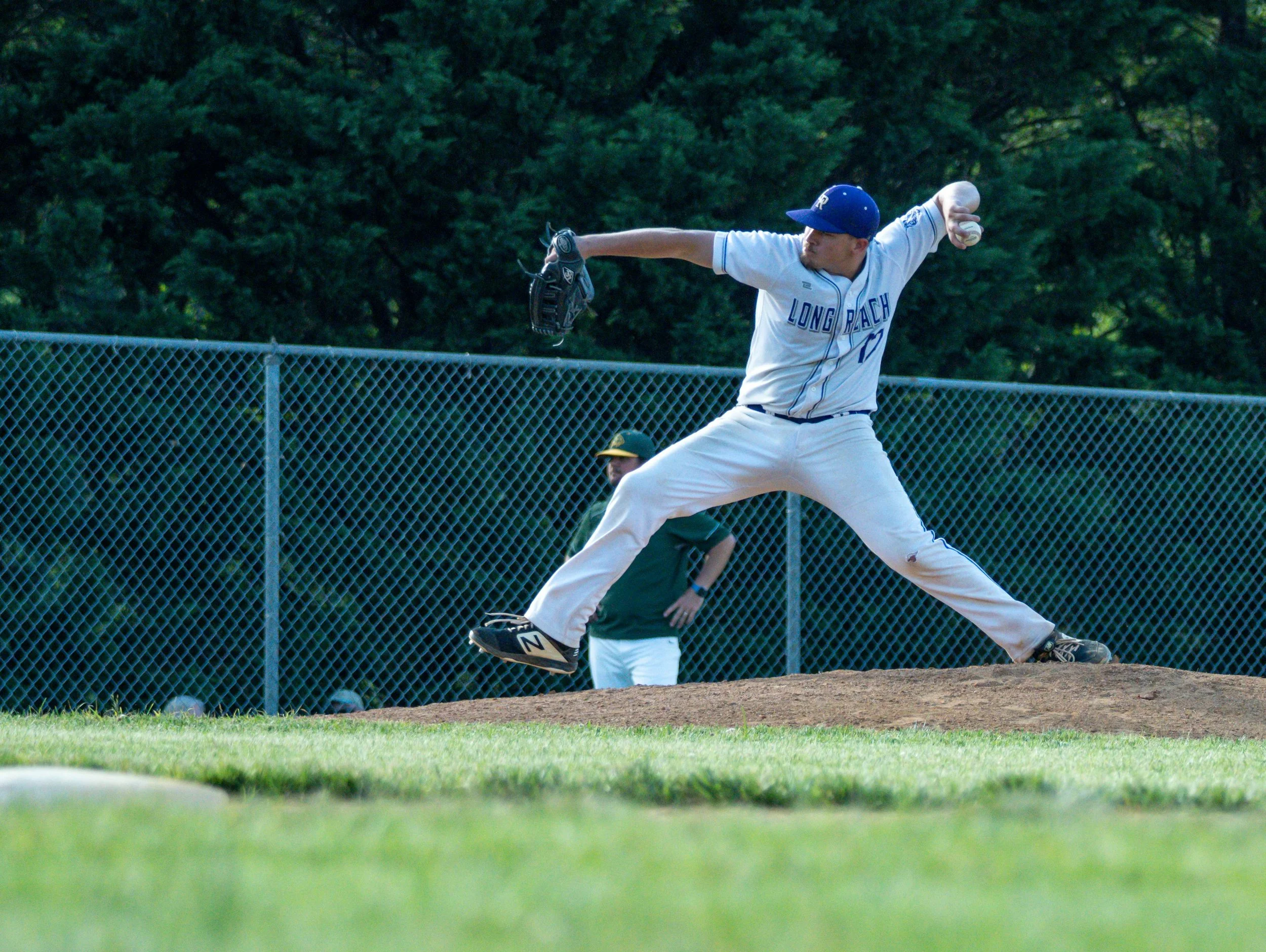 Youth baseball game with three players on the field, one pitcher preparing to throw, in a park with green trees in the background.