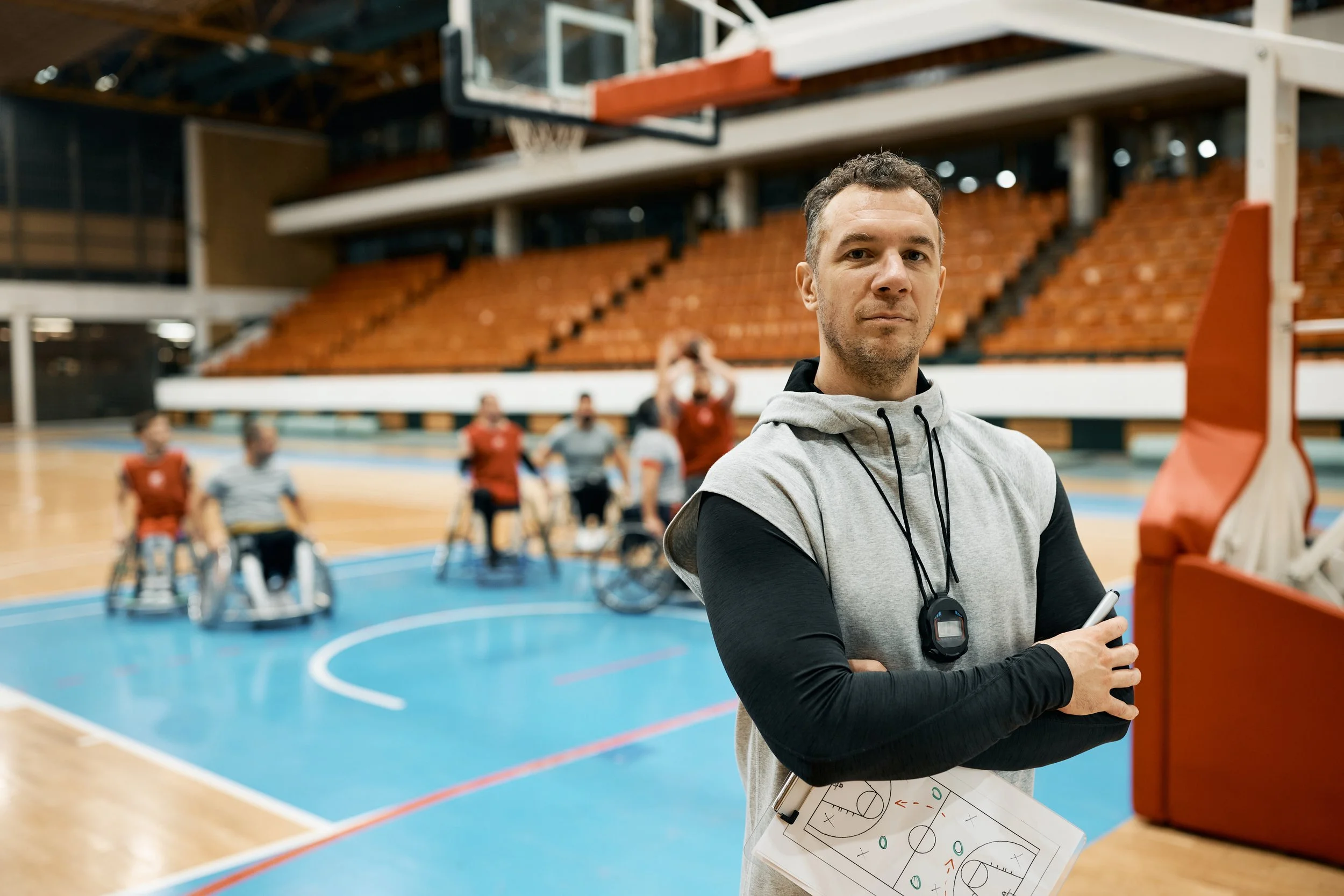 A male coach holding a clipboard and stopwatch on a basketball court with a team of wheelchair basketball players practicing in the background.
