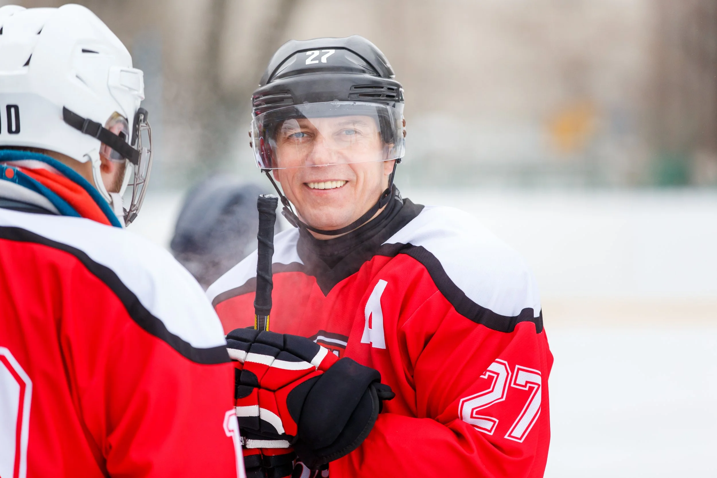 Two ice hockey players in red uniforms, one wearing a helmet with a face shield, are chatting outdoors on the ice rink. One player is smiling, holding a hockey stick.