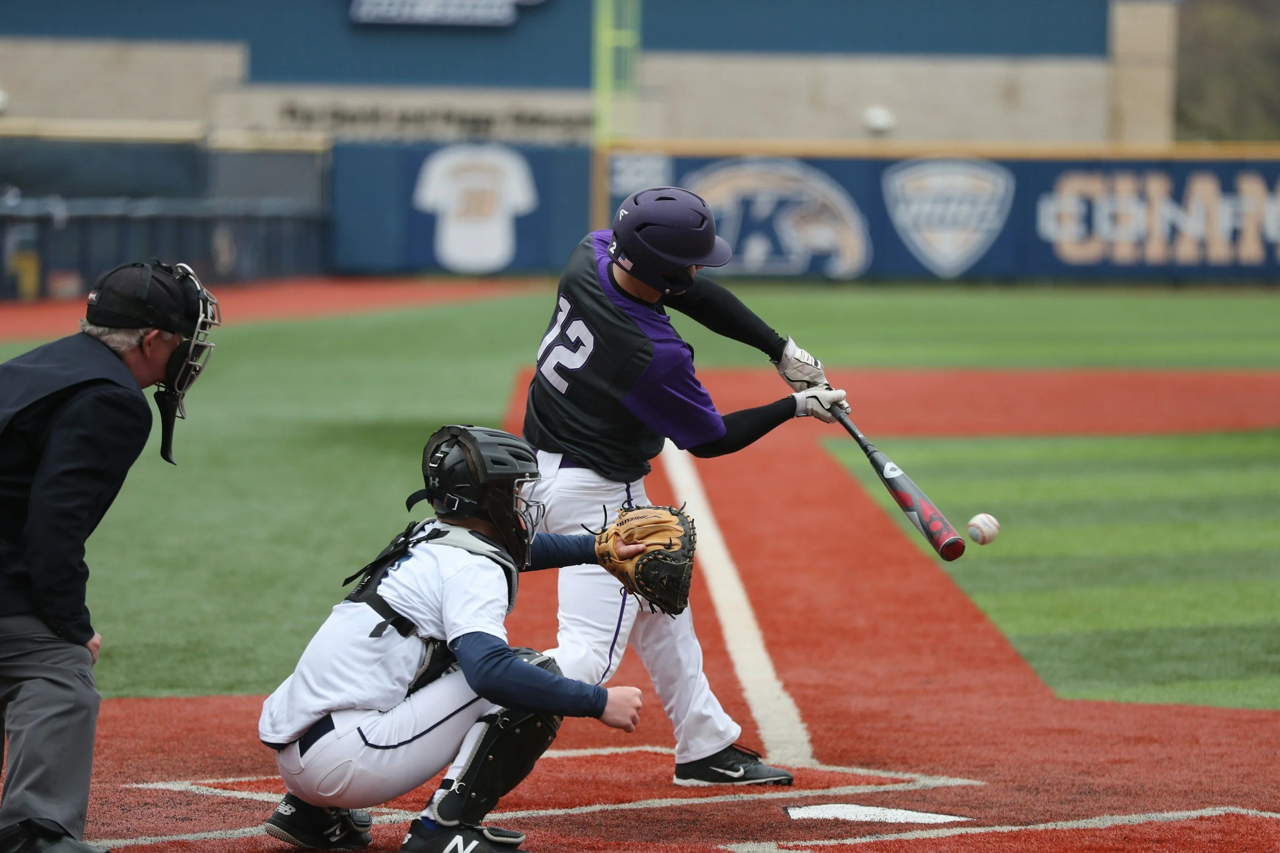 A baseball player in a purple and black uniform swings at a pitched ball during a game on the field, with an umpire and catcher observing.