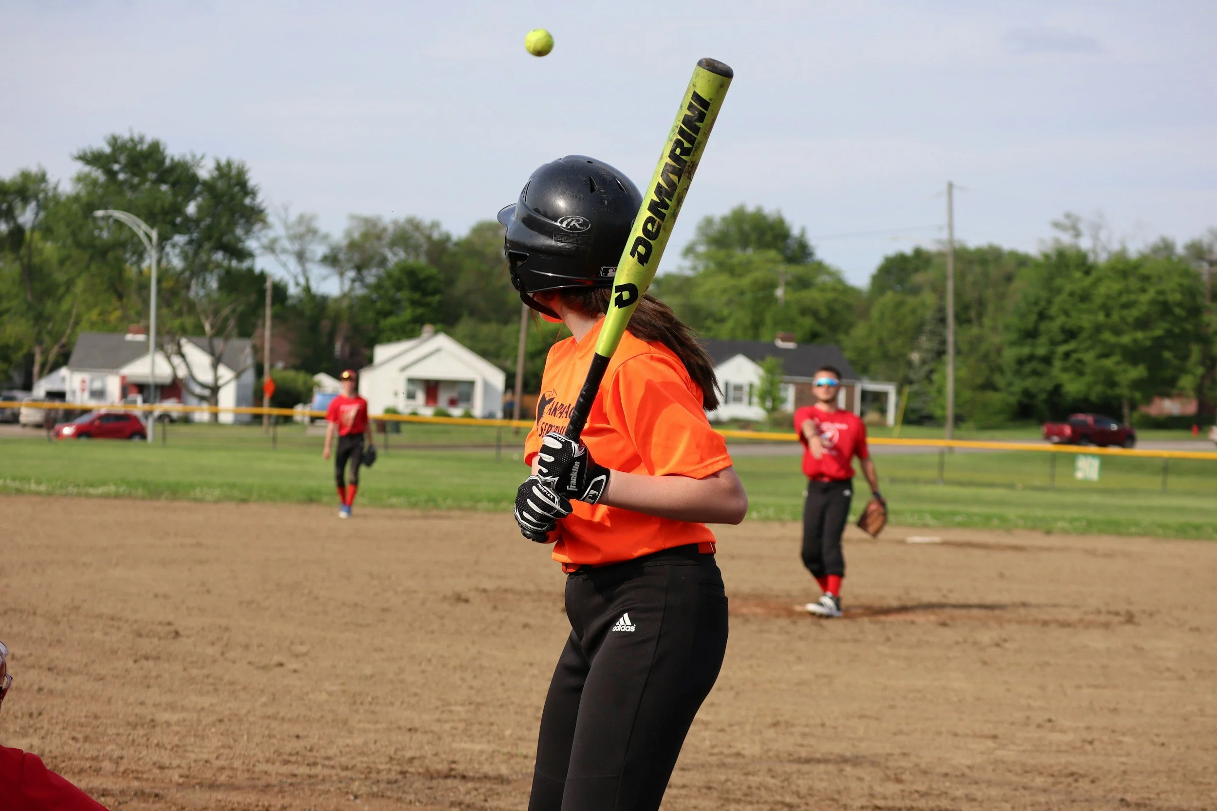 A young softball batter wearing a black helmet, orange jersey, and black pants preparing to hit a ball on a dirt field during a game, with two outfield players and green trees in the background.
