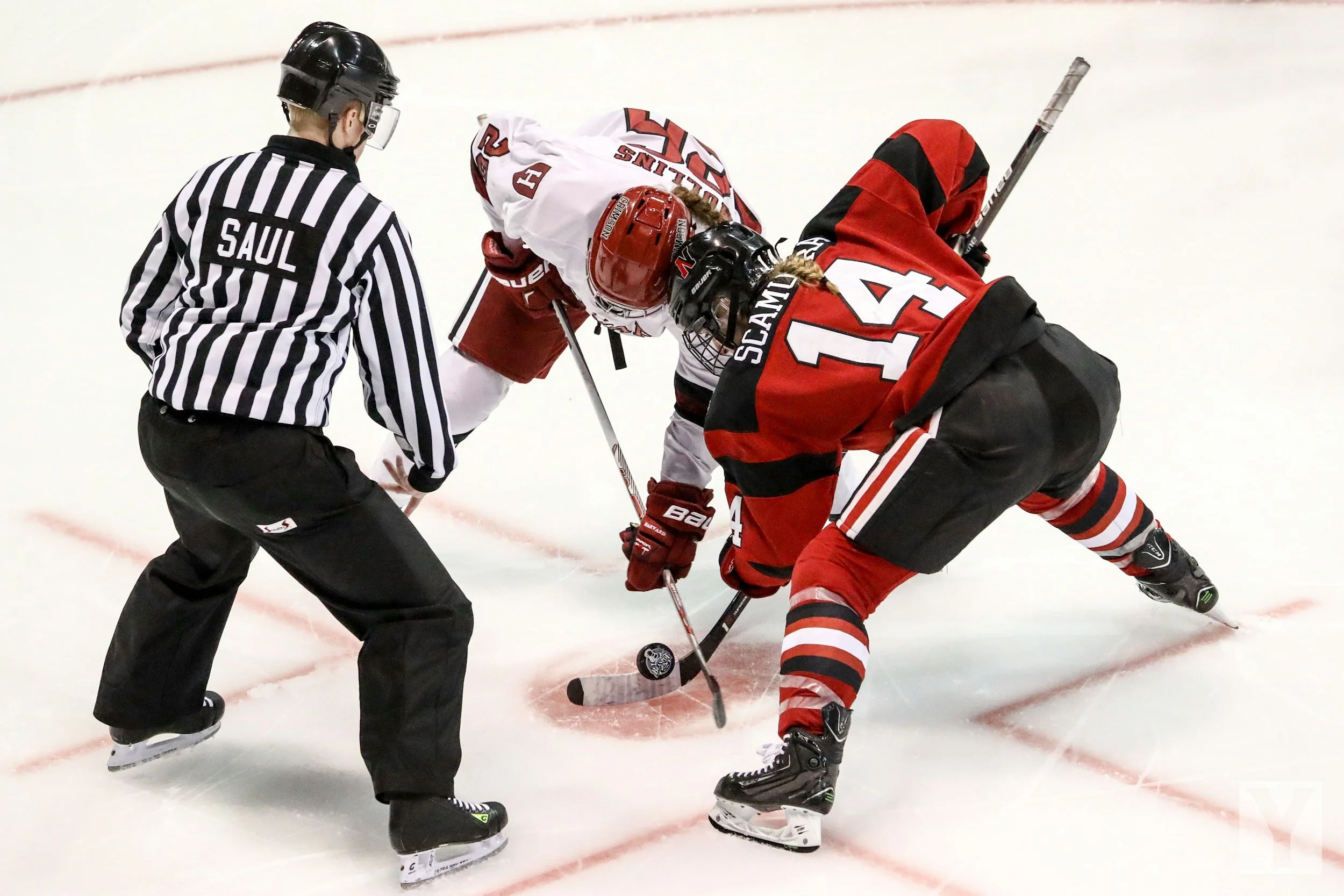 Two ice hockey players from opposing teams face off and compete for control of the puck at center ice, with a referee observing nearby.
