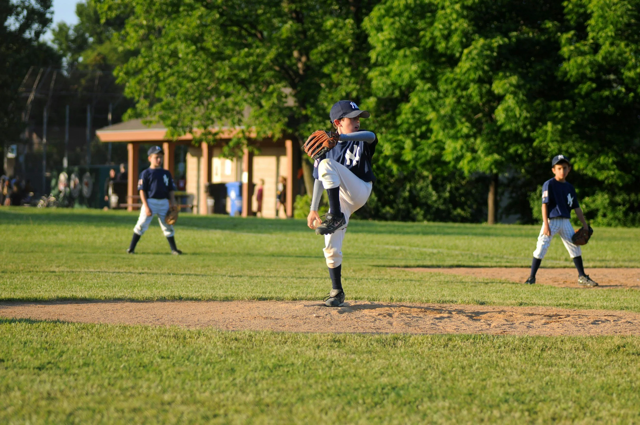 Youth baseball game with three players on the field, one pitcher preparing to throw, in a park with green trees in the background.