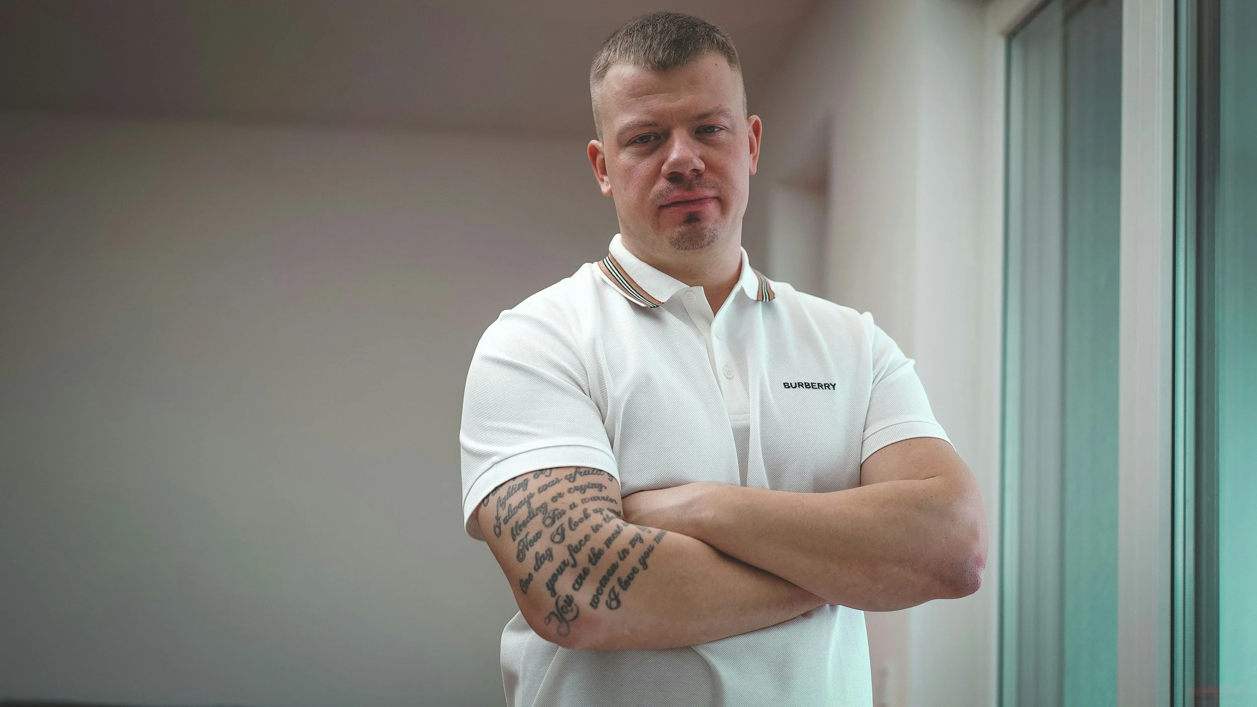A man with short hair and a goatee, standing with arms crossed, wearing a white Burberry polo shirt with a striped collar, indoors near a window with natural light.