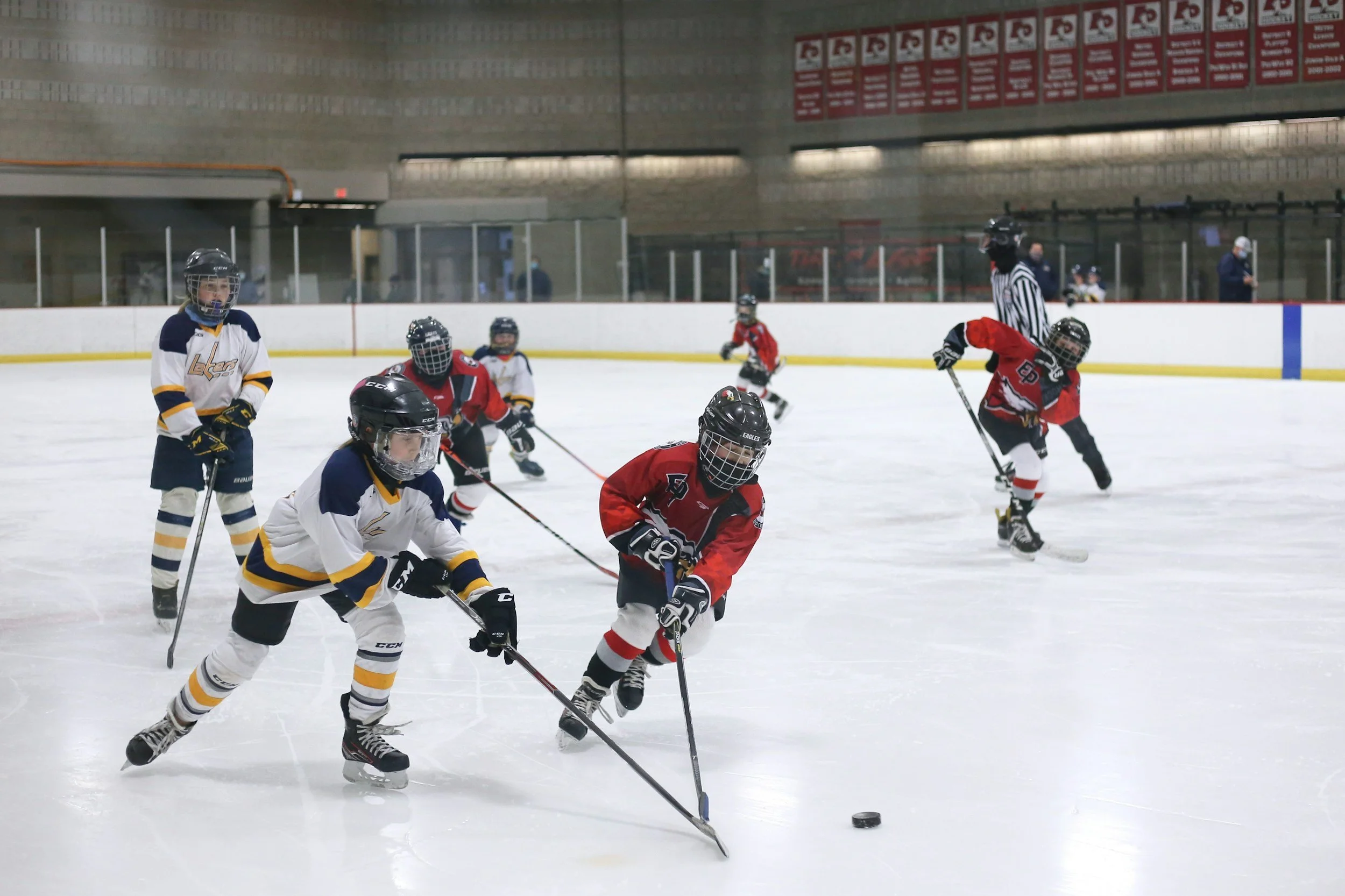 Kids playing hockey on an ice rink with several children in white and yellow jerseys and others in red and black jerseys, wearing helmets and hockey gear, with a referee watching in the background.