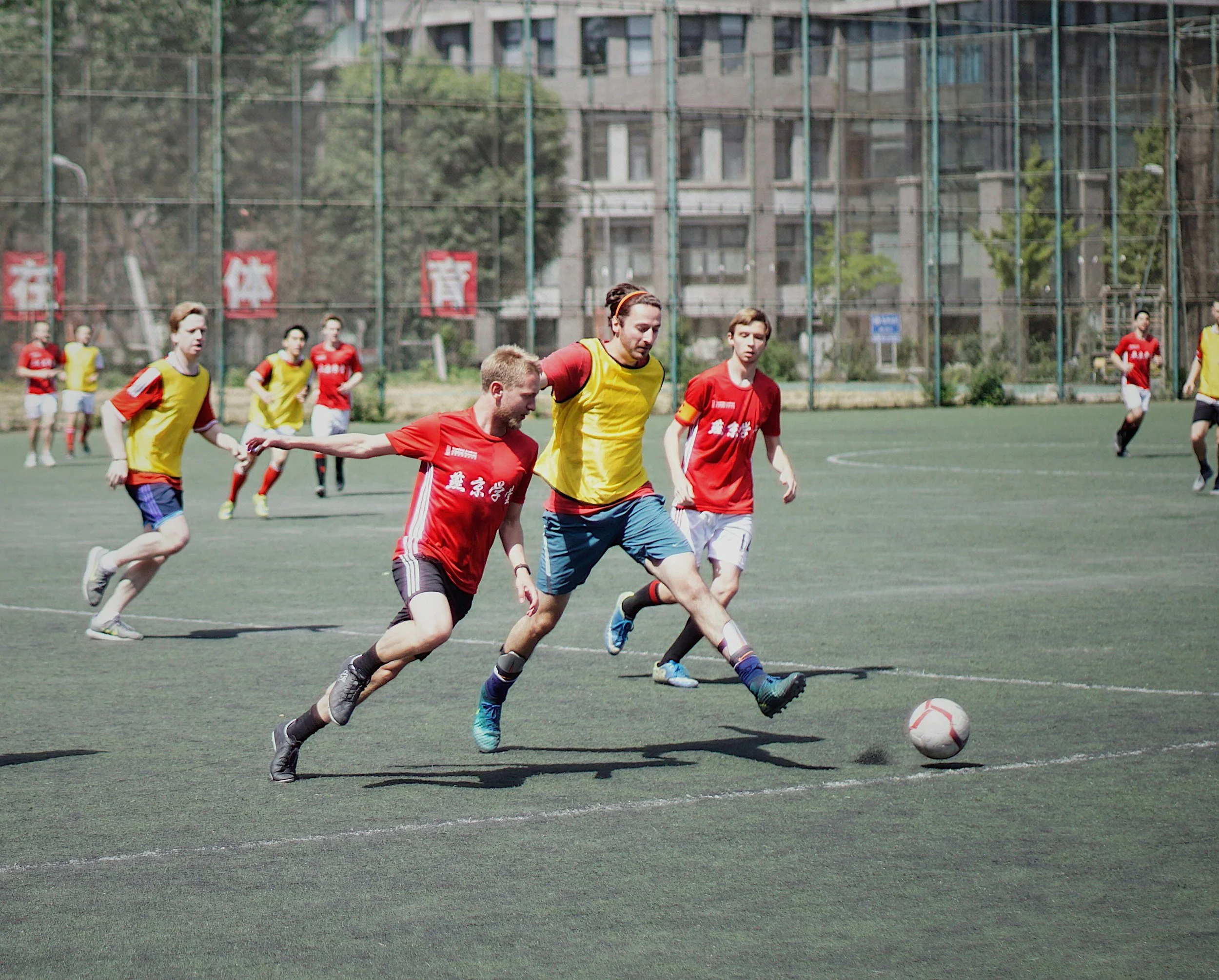 Men playing soccer on a field with a cityscape in the background, wearing red and yellow jerseys.