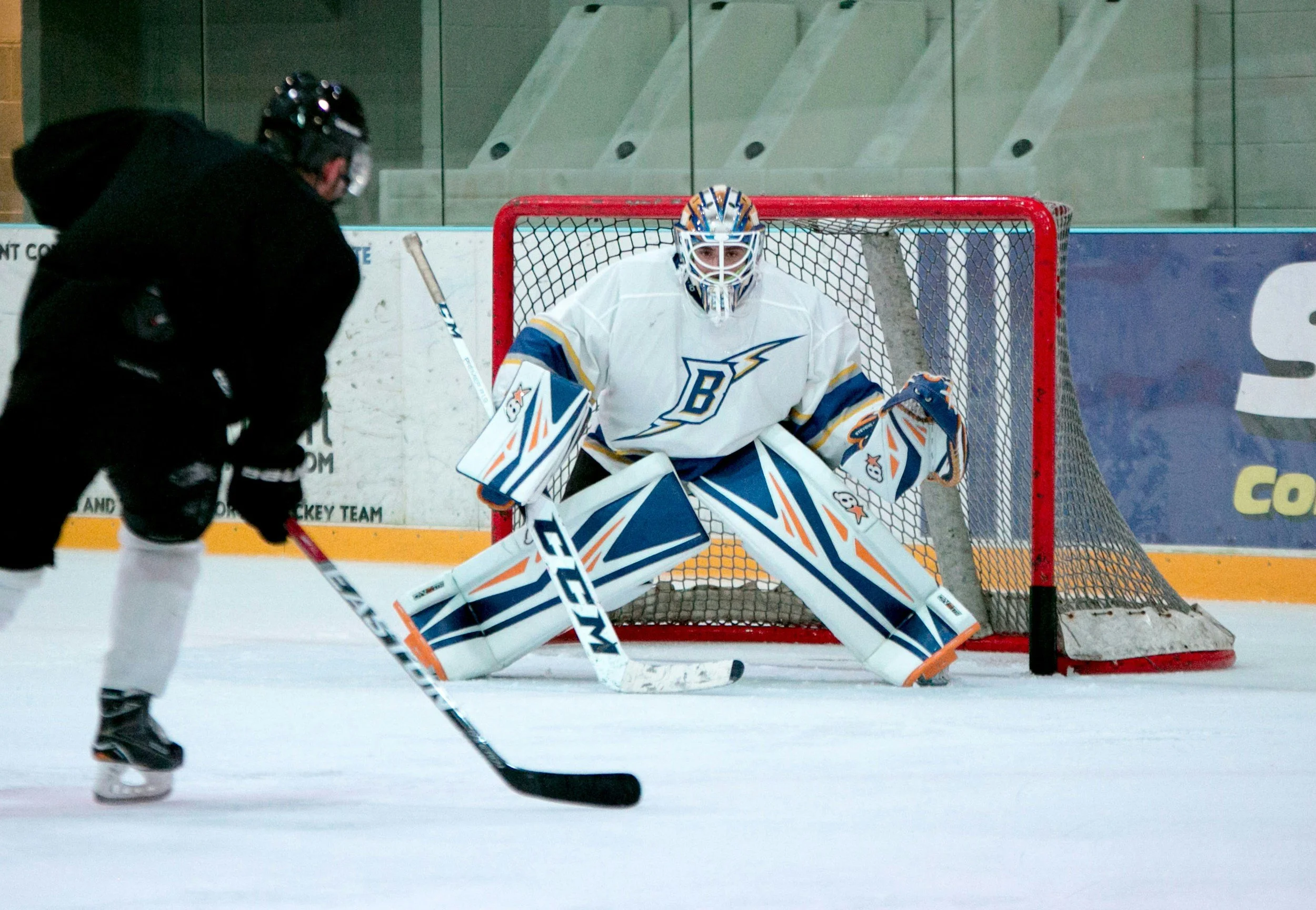 An ice hockey goalie in white, blue, and orange gear prepares to block a shot while a skater in black approaches on the ice during a game.