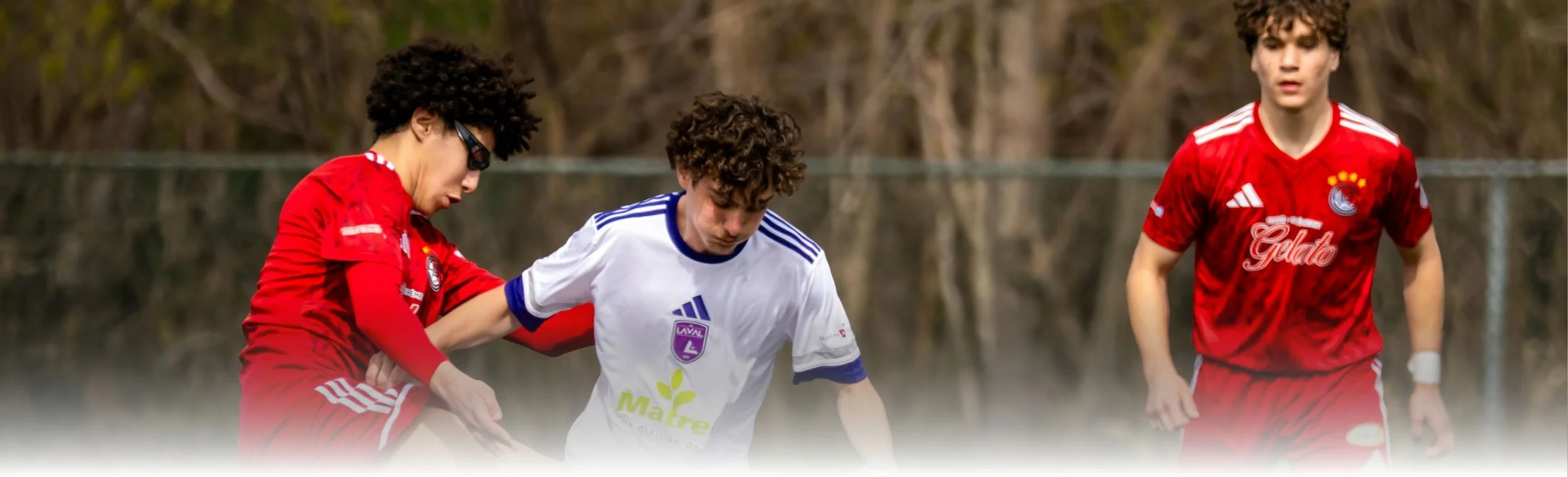 Three young men playing soccer outdoors, with two wearing red jerseys and one in a white jersey, near a body of water with trees in the background.