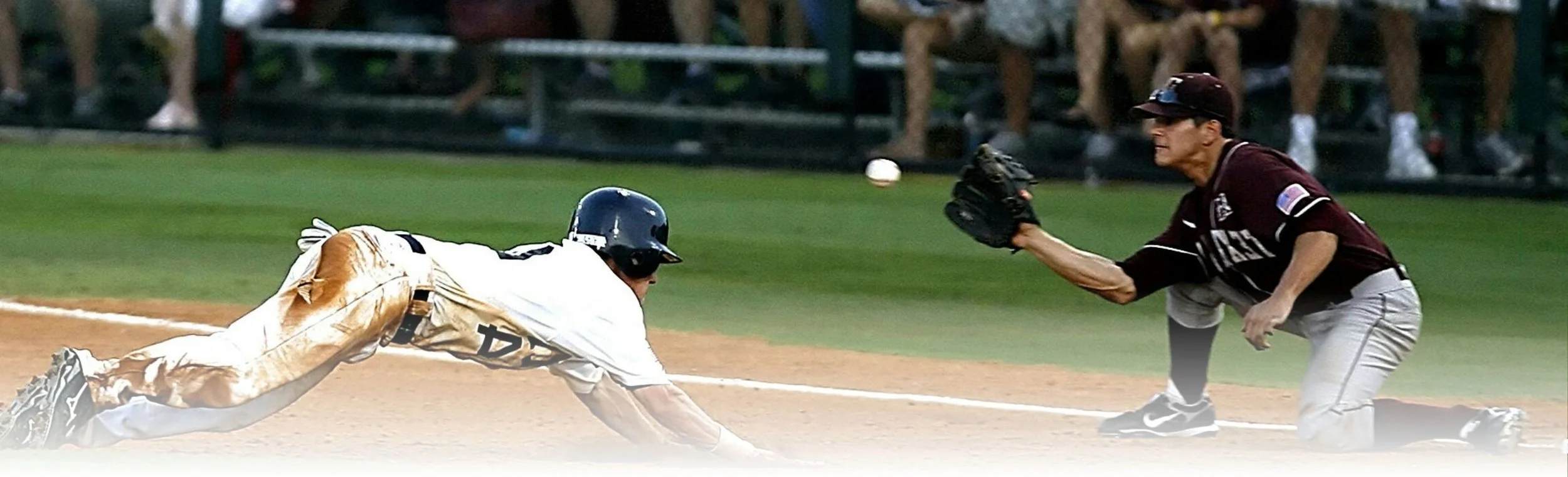 A baseball player in a white uniform is sliding into a base while a player in a maroon jersey reaches out with his glove to catch the ball during a game.