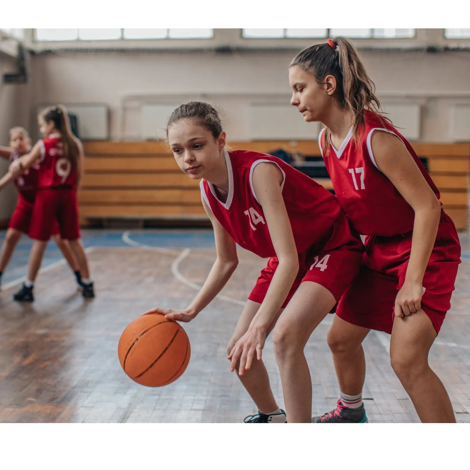 Two young female basketball players in red jerseys and shorts practice dribbling on an indoor court, with a basketball between them.