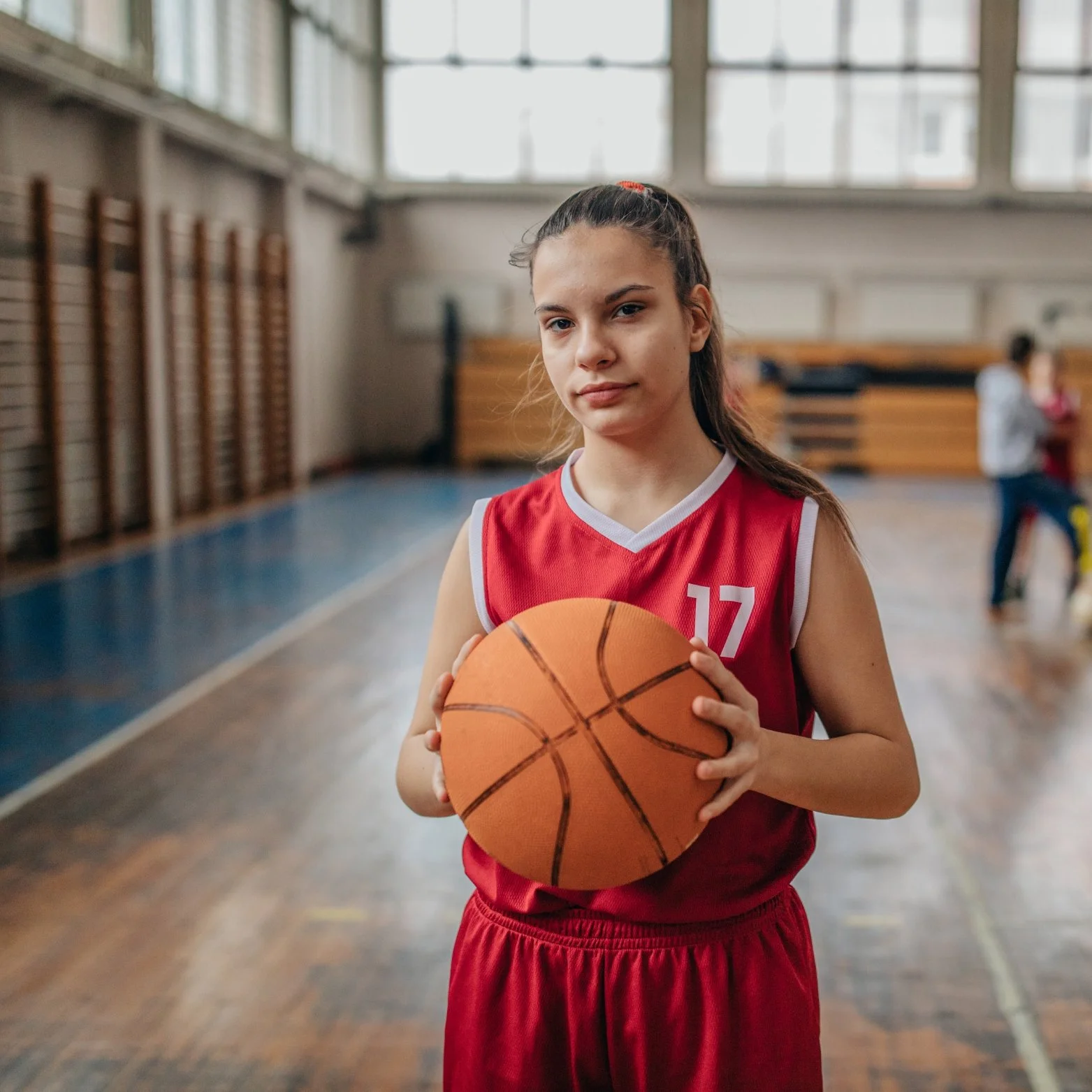 A young girl in a red basketball uniform holding a basketball in a gymnasium.